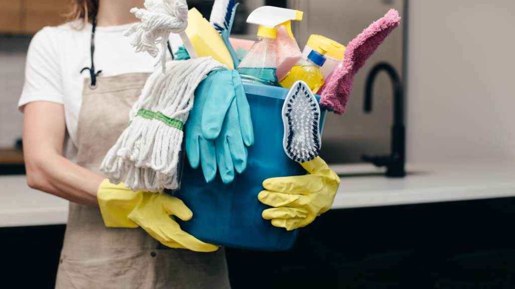 Maid in kitchen in apron and rubber gloves holding bucket of cleaning supplies